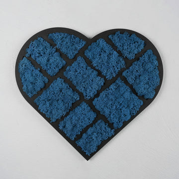 Das Bild zeigt ein herzförmiges Wandkunstwerk, das aus blauem Mooselementen besteht, die in quadratischen Segmenten angeordnet sind. Diese Segmente sind auf einem schwarzen Hintergrund befestigt, was einen starken Kontrast zwischen den blauen und schwarzen Elementen erzeugt. Der Hintergrund ist eine weiße Wand, die das Kunstwerk zusätzlich hervorhebt. Die Textur des Mooses ist detailliert und gut sichtbar.